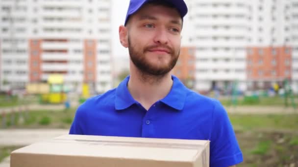 A male courier delivery man with a cardboard box is walking along the street - Footage, Video