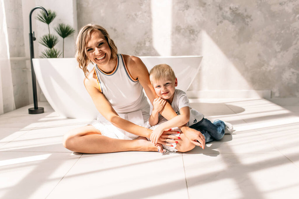 молодая мама, красивая девушка в белой юбке и белой футболке, в просторной светлой ванной комнате, рядом с ней мальчик, он обнимает ее, они счастливы и улыбаются
 - Фото, изображение