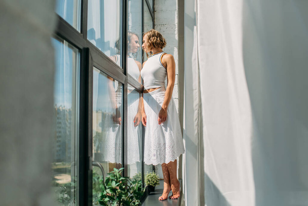 young beautiful girl in a white skirt and white t-shirt, in a spacious bright bathroom, she is on the windowsill near the high window, the bright sun shines on her, she is happy - Photo, image