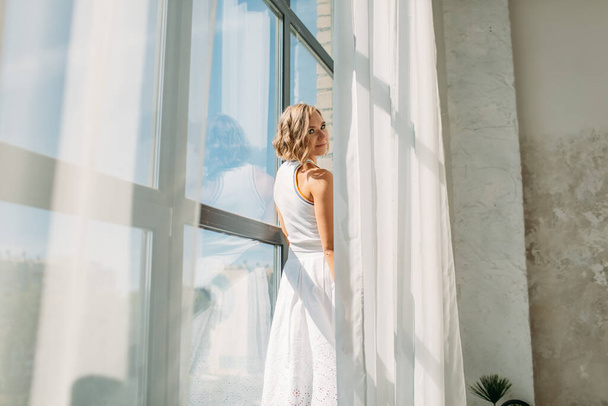 young beautiful girl in a white skirt and white t-shirt, in a spacious bright bathroom, she is on the windowsill near the high window, the bright sun shines on her, she is happy - Фото, изображение
