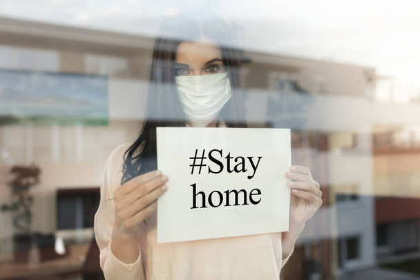 Young woman with warning paper sign looking through window, home isolation concept. Covid-19 pandemic situation in self isolation at home. New corona virus changes normal lives. - Photo, Image