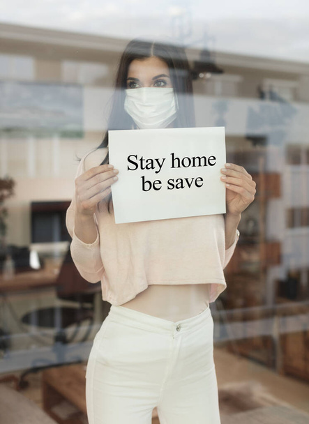 Młoda kobieta ze znakiem ostrzegawczym patrząca przez okno, koncepcja izolacji domu. Pandemia Covid-19 w izolacji domowej. Nowy wirus korony zmienia normalne życie.. - Zdjęcie, obraz