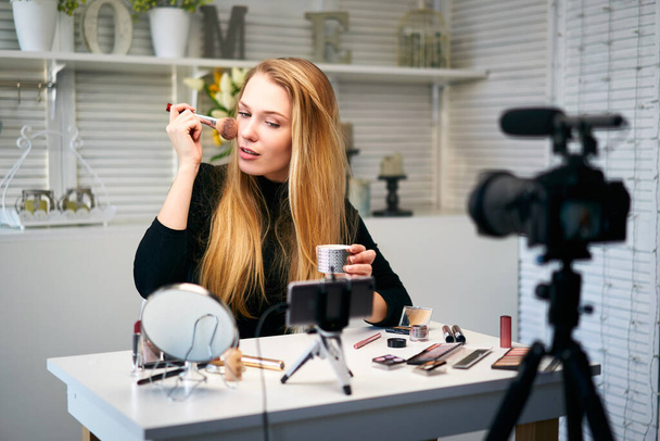 Beauty blogger woman filming daily make-up routine tutorial at camera on tripod. Influencer girl live streaming cosmetics product review in home studio. Vlogger female applies skin powder with brush. - Foto, Imagen