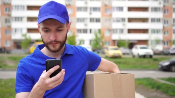 Ein männlicher Kurierfahrer hält eine Schachtel in der Hand und benutzt ein Smartphone - Filmmaterial, Video