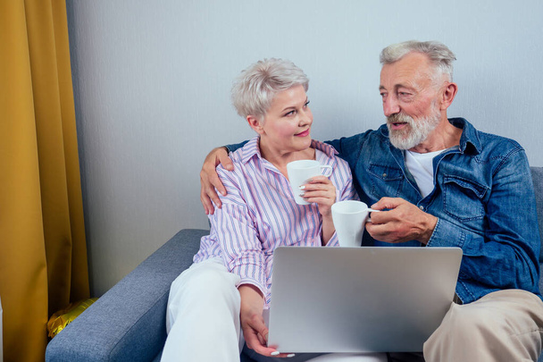 elderly couple in love sitting gray couch in living room look at screen laptop and drinking cup of tea or coffee - Foto, Bild