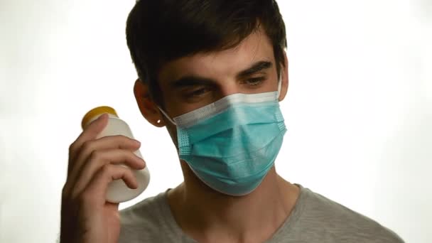 Portrait of a young man with a medical mask who shakes a can of pills and looks at it on a white background - Felvétel, videó