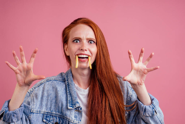 funny redhaired ginger woman with potatoes like canine tooth scaring, her hands want grab you in studio pink background. fright candies or life concept - Fotografie, Obrázek
