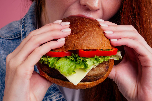 cropped photo of redhaired ginger redhead woman eating big cheeseburger with cheese,beef cutlet and tomato lettuce and licking fingers close up - Φωτογραφία, εικόνα