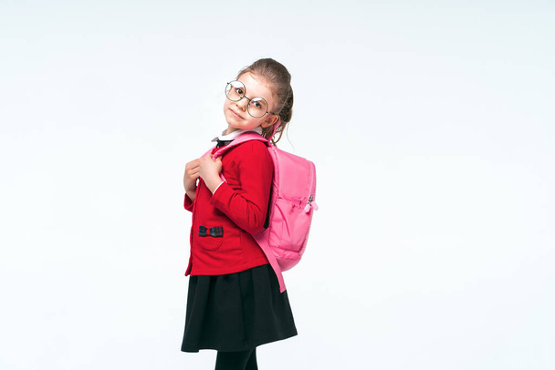 Αξιολάτρευτο κοριτσάκι με κόκκινο σχολικό μπουφάν, μαύρο φόρεμα, στρογγυλά γυαλιά που κρατιούνται από τα λουριά ενός σακιδίου και χαμογελούν και κοιτάζουν την κάμερα, ποζάροντας σε λευκό φόντο στούντιο. Απομόνωση - Φωτογραφία, εικόνα