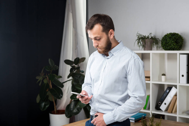 Theme office business. Handsome young caucasian man confident and strong with beard standing in bright room on working place. dressed stylishly shirt and trousers. Uses technology. Hand mobile phone. - Photo, Image