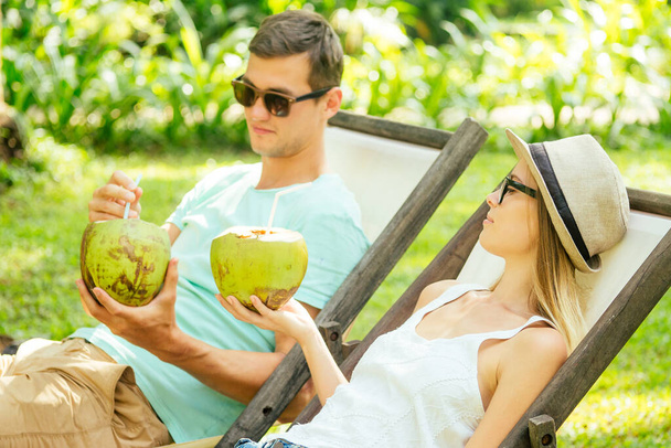 Young loving couple drink coconut while sitting under palms in deck chair lounge resort - Fotoğraf, Görsel
