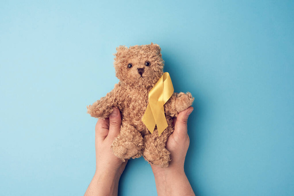 a női kezek egy sárga szalaggal összehajtogatott kis játékmackót tartanak egy kék háttérben. a gyermekkori rák elleni küzdelem koncepciója. öngyilkosságok problémája - Fotó, kép