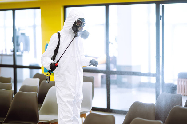 Man wearing protective suit disinfecting assembly hall with spray chemicals to preventing the spread of coronavirus, pandemic in quarantine city. Disinfecting of office. COVID-19. - Photo, image