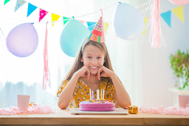 Blank meisje lacht dromerig en kijkt naar verjaardagsregenboogtaart. Feestelijke kleurrijke achtergrond met ballonnen. Verjaardagsfeest en wensenconcept. - Foto, afbeelding