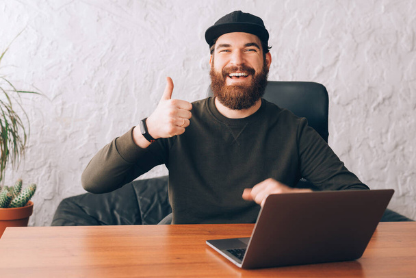 счастливый улыбчивый офисный работник, сидящий на столе и показывающий жест вверх пальцами
 - Фото, изображение