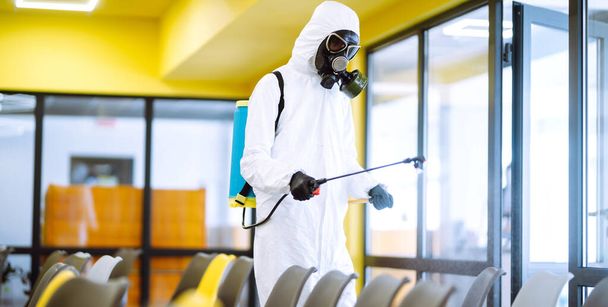 コロナウイルスの拡散を防ぐためにスプレー化学薬品で組立ホールを消毒保護スーツを身に着けている男,検疫都市のパンデミック.オフィスの消毒。COVID-19. - 写真・画像