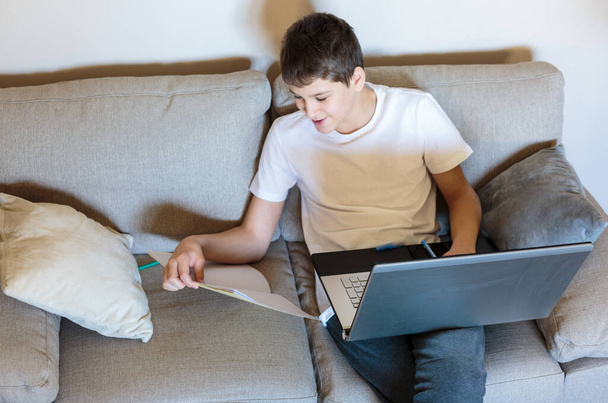 Cute young boy in glasses and white t shirt sitting on the couch in the living room with laptop and study. Homeschooling, self education, distance learning by  kids. - Valokuva, kuva