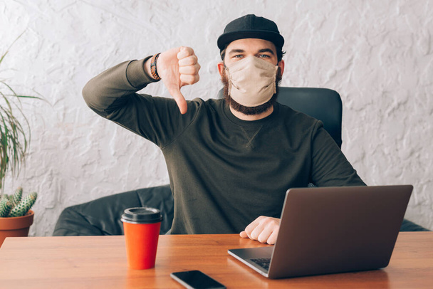 Büroangestellter steht mit Anti-Viren-Maske vor Laptop und zeigt Daumen nach unten - Foto, Bild