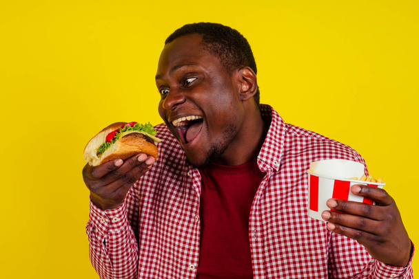 handsome latin man eating fries nuggets and burger with appetite and enjoyment in studio yellow background.chicken legs in a bucket - Foto, Imagem