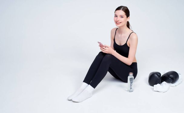 Image of a sporty pretty young Caucasian woman, in a black top and leggings, sitting on a white background, resting after a workout, holding a smartphone in her hands, smiling and looking straight. - Foto, Imagem