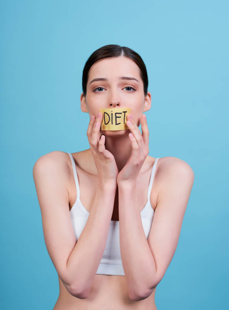Young sad emaciated woman in a white top, with a taped mouth with the word diet, stands on a blue background. Hungry young woman with a closed mouth. Healthy lifestyle concept. Weight loss problem. - Foto, Bild