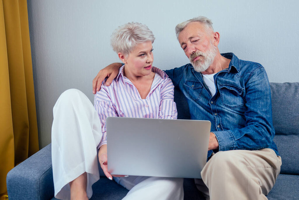 elderly couple in love sitting gray couch in living room look at screen laptop - Foto, Bild