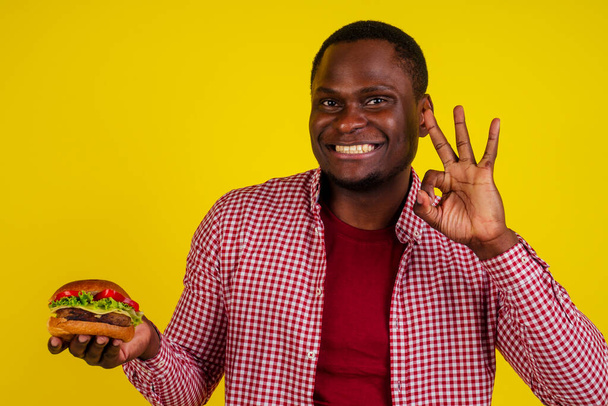 young african american man eating hamburger isolated on yellow background showing ok gesture. - Foto, afbeelding