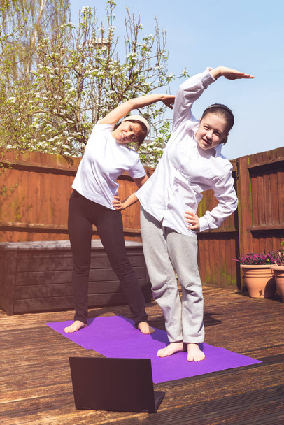 Glückliche Mutter und Tochter machen Online-Yoga mit Laptop während der Selbstisolierung im Garten, kein Gerätetraining, Meditationstipps für Anfänger, zu Hause bleiben und gesund bleiben Konzept - Foto, Bild