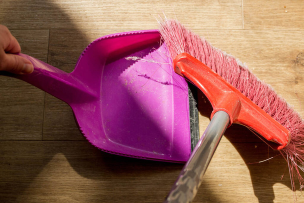 Tools for cleaning the house. A red sweeping brush with artificial bristles and a long handle and a purple dustpan against the background of the vinyl floor covering in the kitchen close up during cleaning - Photo, Image