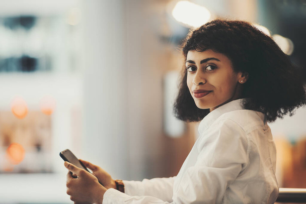 An indoor portrait of a young dazzling African-American woman in a white shirt with a cellphone and curly Afro hair looking at camera; a charming biracial woman entrepreneur with a smartphone in hands - Photo, image