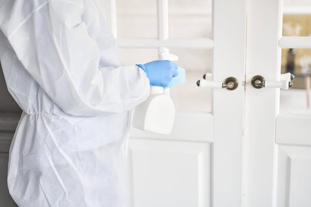 Disinfezione, pulizia e lavaggio delle maniglie delle porte. COVID-19. Prevenzione dell'infezione da coronavirus. Una donna in tuta protettiva e guanti blu spruzza una soluzione disinfettante
. - Foto, immagini