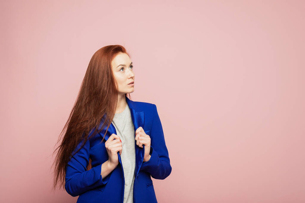 A redhead girl is on the pink background. A thoughtful redhead girl is on the pink background. A girl is in blue jacket on the pink background. A girl with beautifull red hair on the pink background. A girl is looking into the distance. - Photo, Image
