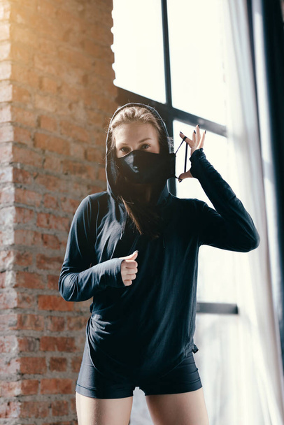 A young girl in a black hoodie corrects the black mask on the face. Boxing coach took a break during a home workout. - Foto, Imagen