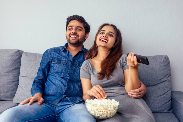 young indian couple watching comedy movie with remote control in hand in living room with popcorn - 写真・画像