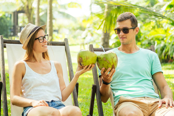 Young loving couple drink coconut while sitting under palms in deck chair lounge resort - Foto, Imagem