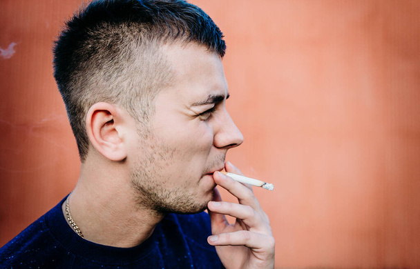 Portrait of a young man smoking a cigarette near a building wall - 写真・画像