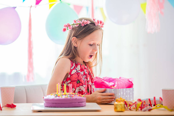 Biała dziewczyna uśmiecha się i patrzy na urodzinowy tort tęczowy. Uroczyste kolorowe tło z balonami. Urodziny i koncepcja życzeń. - Zdjęcie, obraz