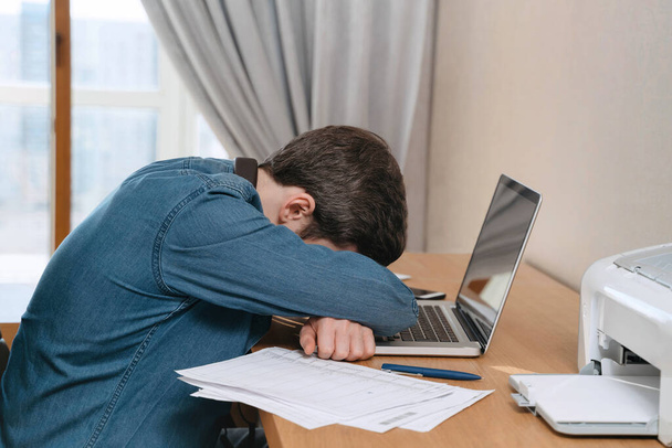 Burnout jongeman huilt in zijn kantoor, zit aan zijn bureau met een laptop en documenten. Moe depressieve freelancer gefrustreerd door zakelijk falen, voel me uitgeput, bezorgd over werkproblemen - Foto, afbeelding