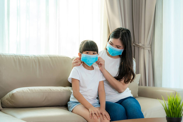 Aasialainen äiti yllään tyttärensä terve kasvonaamio istuu olohuoneessa kotona estää PM2,5 pöly, savusumu, ilmansaasteet ja COVID-19. Terveydenhuollon käsite. - Valokuva, kuva