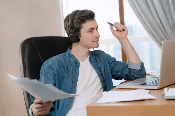 Ευτυχισμένος τύπος φορώντας ακουστικά απολαμβάνοντας την εξ αποστάσεως εκπαίδευση του, κάθεται στο γραφείο του και κοιτάζοντας οθόνη laptop, βλέποντας webinar ακούγοντας σε απευθείας σύνδεση πορεία, εκμάθηση γλώσσας, μελέτη με δάσκαλο - Φωτογραφία, εικόνα