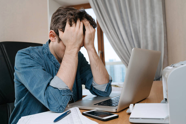 Väsynyt stressaantunut nuori mies istuu kannettavan tietokoneen edessä, turhautunut ongelmiin tai tutkimuksen epäonnistumiseen, uupunut, päänsärky, järkyttynyt huonoista uutisista, rahaongelmasta, epäonnistuneesta tentistä tai testituloksesta
 - Valokuva, kuva