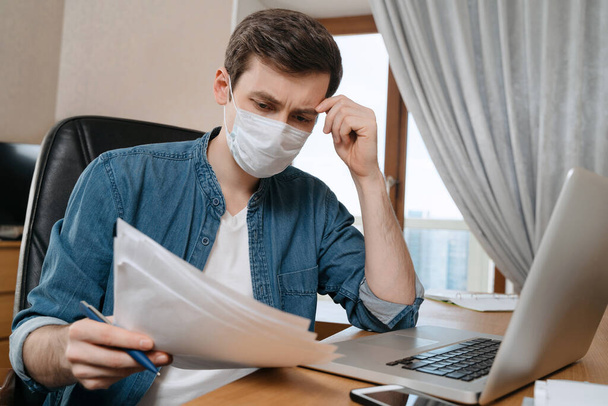 Young thoughtful man in protective face mask studying and working remotely due to Coronavirus or COVID-19 quarantine. Online training education and freelance work with laptop from home. Closing schools, lost job - Fotó, kép
