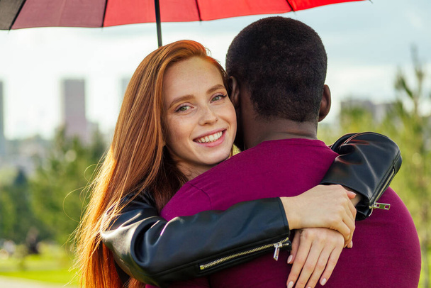 redhaired ginger woman and african american man standing close to each other under umbrella in summer park - Fotografie, Obrázek