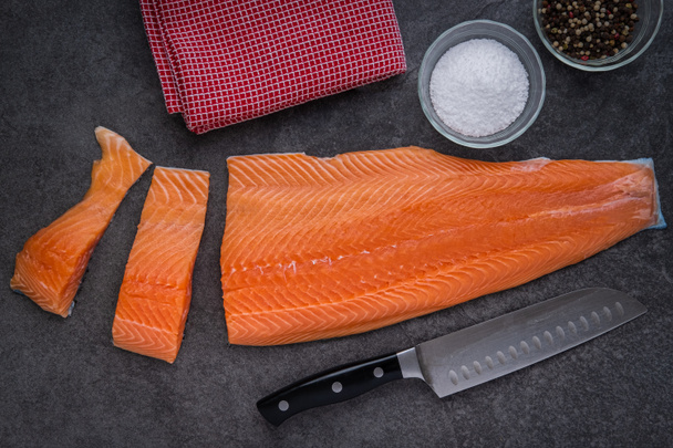 Filetes de salmón cortados con un cuchillo Santoku, sal y pimienta y toalla de cocina en una encimera de pizarra
 - Foto, Imagen