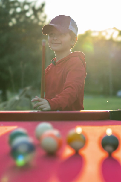 Ένα μικρό αγόρι παίζει μπιλιάρδο σε ένα υπαίθριο περιβάλλον - Φωτογραφία, εικόνα