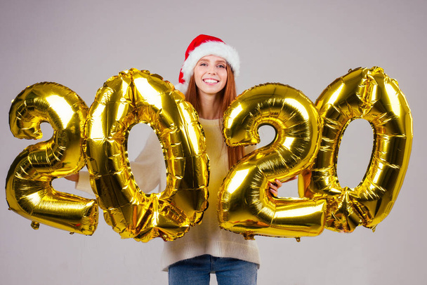 beautiful redhaired ginger woman wearing white knitted sweater and santa claus hat holding gold ballons 2020 numbers in studio background - Fotografie, Obrázek