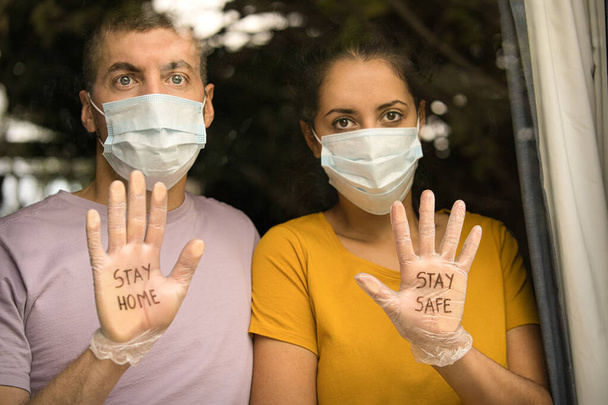 顔のマスクと衛生的な手袋を身に着けているカップルは、家にいて、安全にしてください。家の窓から外を見てる。コロナウイルスと検疫の概念 - 写真・画像