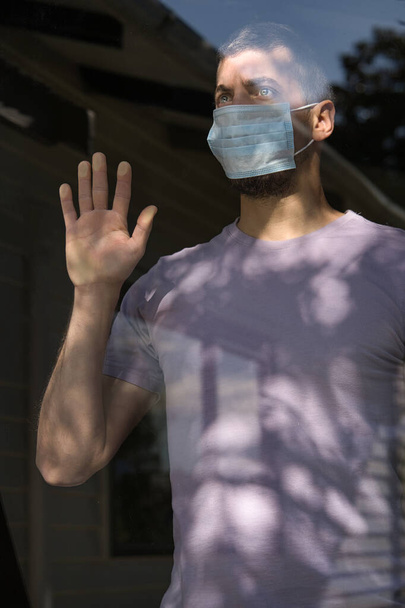 Kaukasischer junger Mann mit Gesichtsmaske, der aus dem Fenster schaut, zu Hause isoliert. COVID-19 und Quarentine-Konzept. Vertikal - Foto, Bild