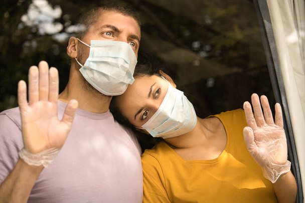Νεαρό ζευγάρι που φοράει μάσκα προσώπου και γάντια υγιεινής μπροστά από το παράθυρο και αγκαλιάζονται. Ιός του Coronavirus και Καραντίνα - Φωτογραφία, εικόνα