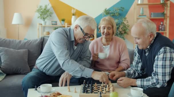 Male friends enjoying chess game while elderly woman drinking tea in apartment - Záběry, video